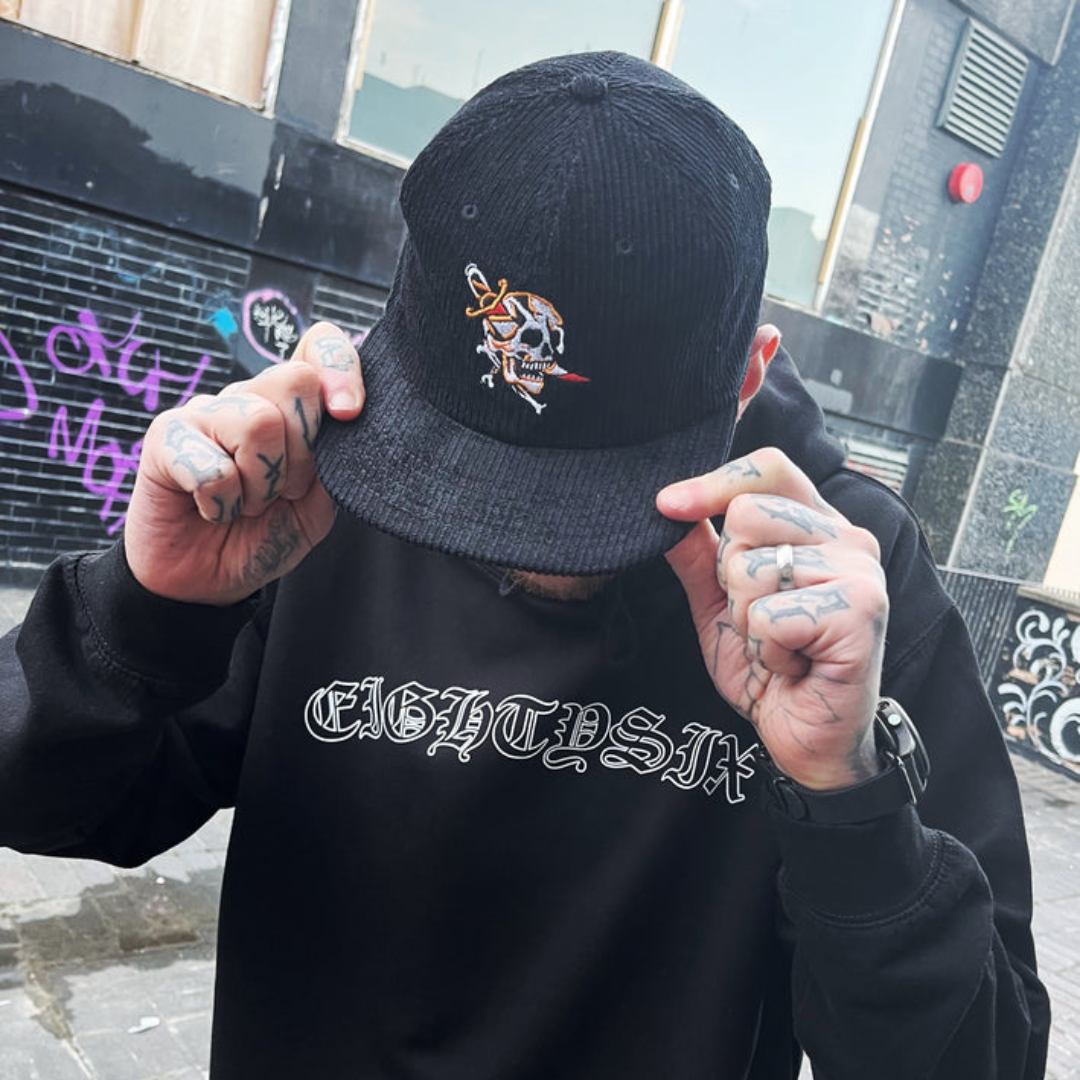 Dagger skull cord cap in black worn by a tattooed model, design features a traditional tattoo style skull with a dagger through its cheek