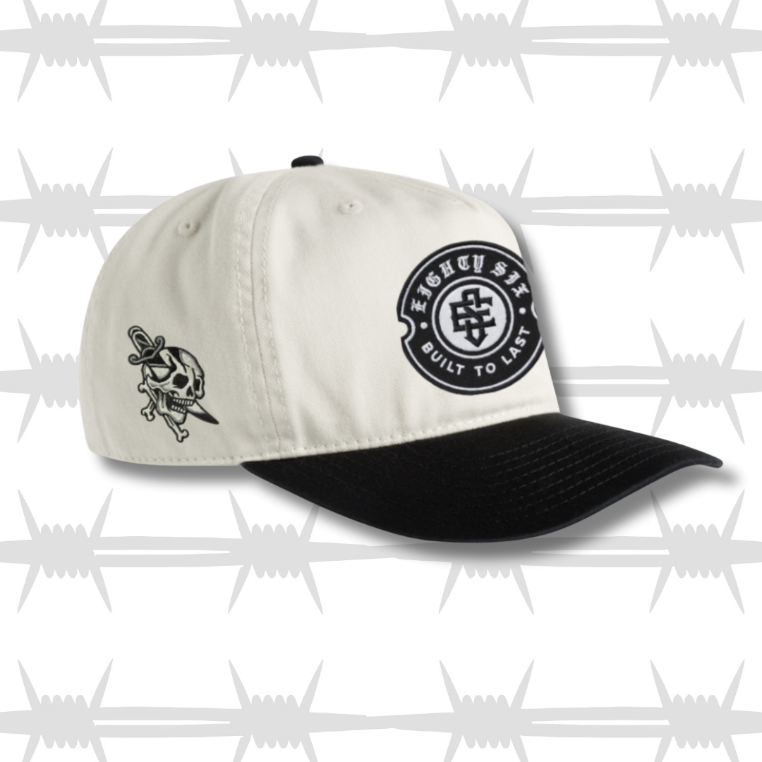 White and black two tone cap with black embroidered tattoo inspired skull and dagger design and built to last crest logo
