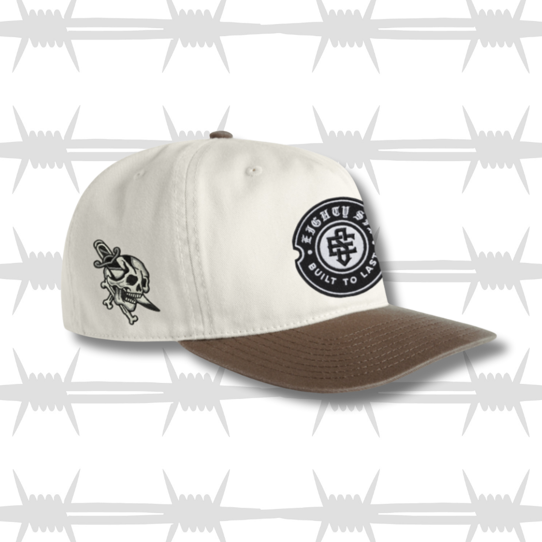 White and brown two tone cap with black embroidered tattoo inspired skull and dagger design and built to last crest logo