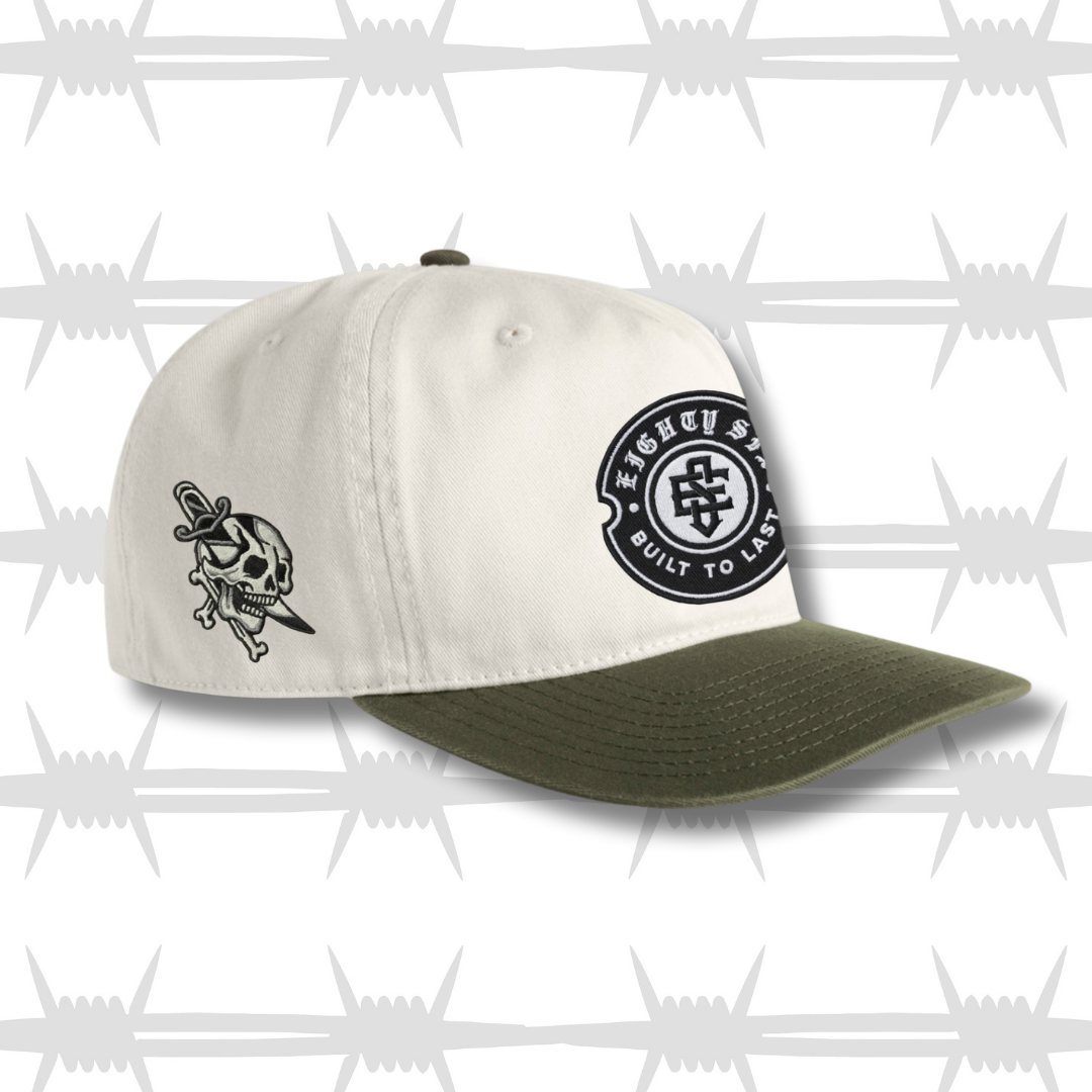 White and green two tone cap with black embroidered tattoo inspired skull and dagger design and built to last crest logo
