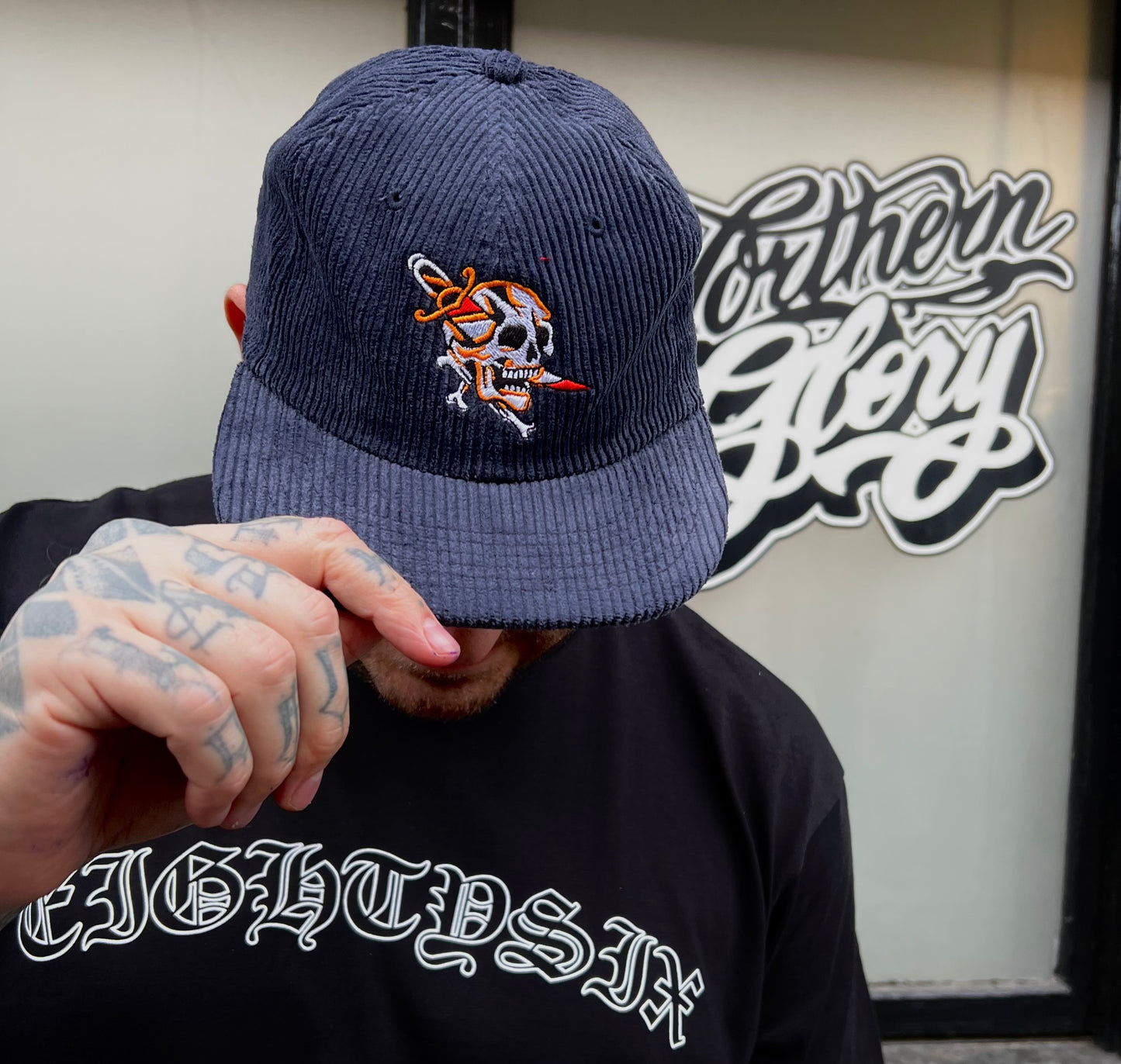 Dagger skull cord cap in blue worn by a tattooed model, shows a traditional style tattoo skull with dagger design embroidered on the front centre of a corduroy cap.